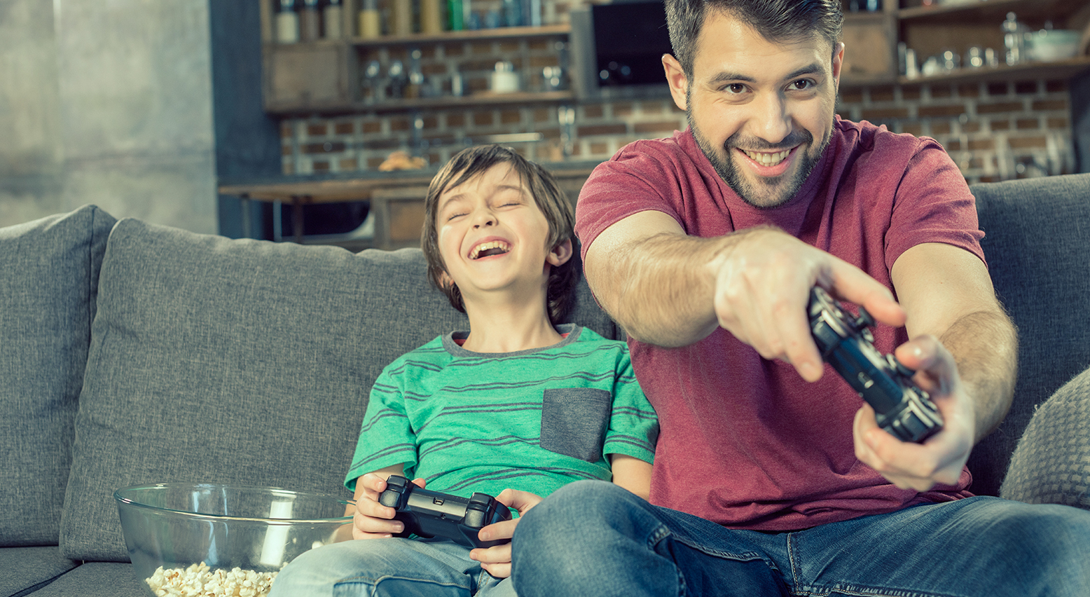 Junge und Vater sitzen auf der Couch mit Controllern in der Hand, gemeinsam ein Videospiel spielend