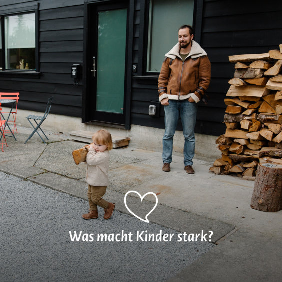 Kleinkind trägt Holzscheit und Vater guckt ihm dabei zu.