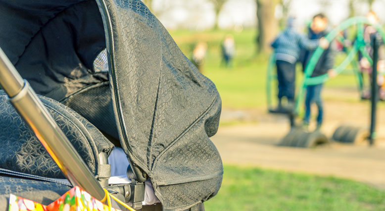 Bild: im Vordergrund ein Kinderwagen, im Hintergrund ein Spielplatz.