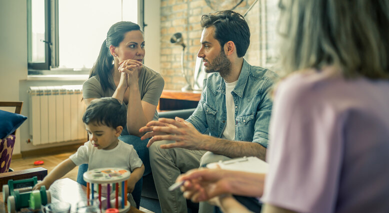 Ein Elternpaar spricht mit einer Beraterin, während ein Kleinkind am Tisch spielt. 